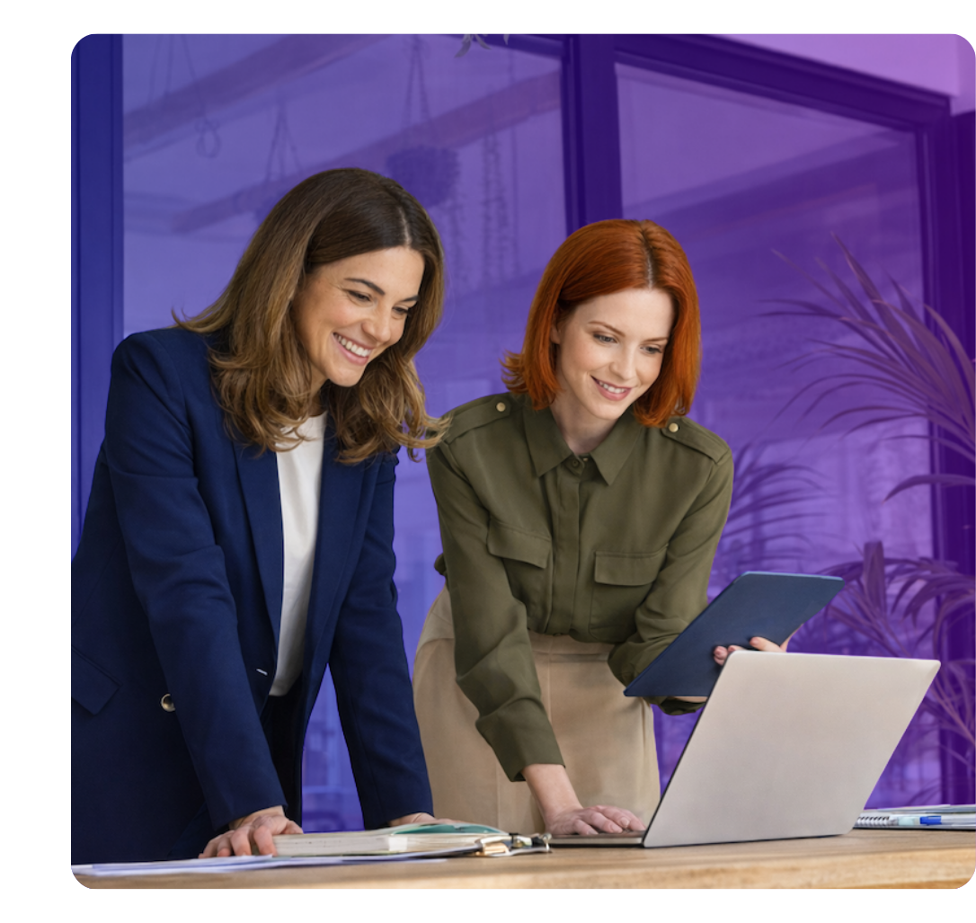 Two colleagues collaborate over a laptop in a modern office, smiling as they review information together, with one holding a tablet and the other referencing documents on the desk.